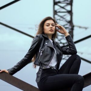 Confident woman in black leather jacket poses on industrial structure under overcast sky.